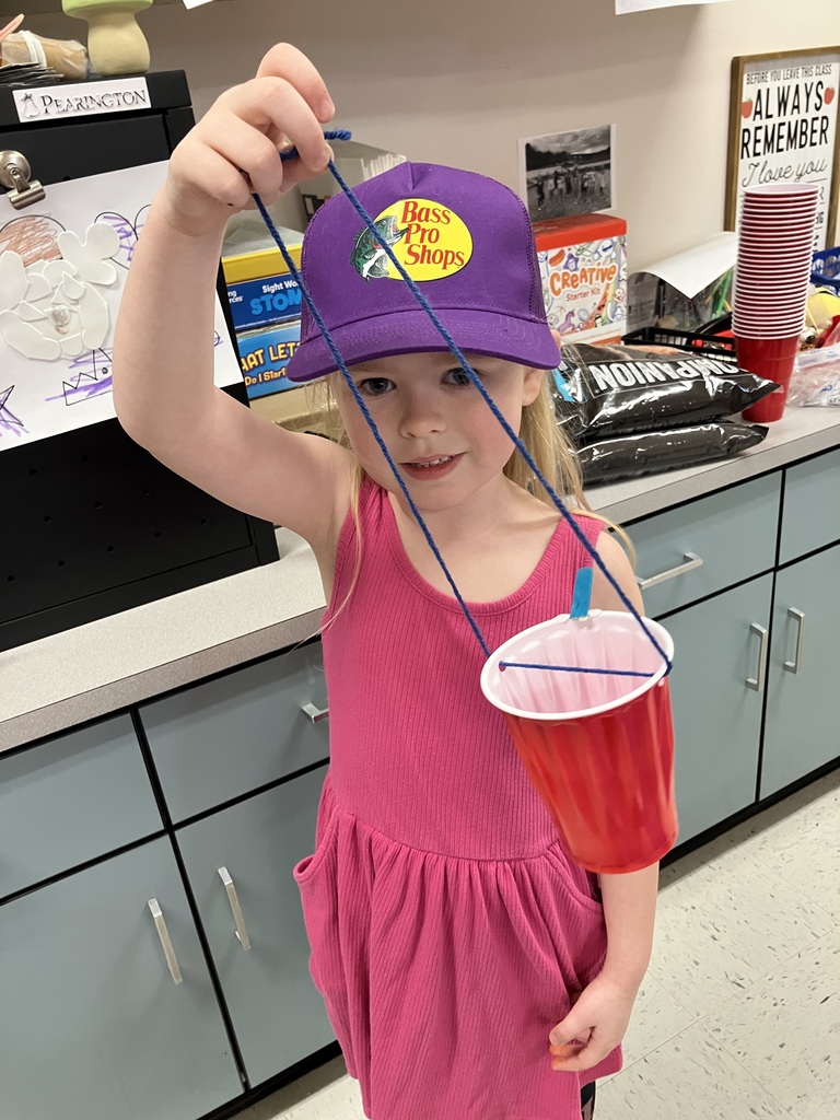  A young girl wearing a purple "Bass Pro Shops" hat and a pink dress holds up a red plastic cup by a long blue string. She is standing in front of a gray classroom counter. 