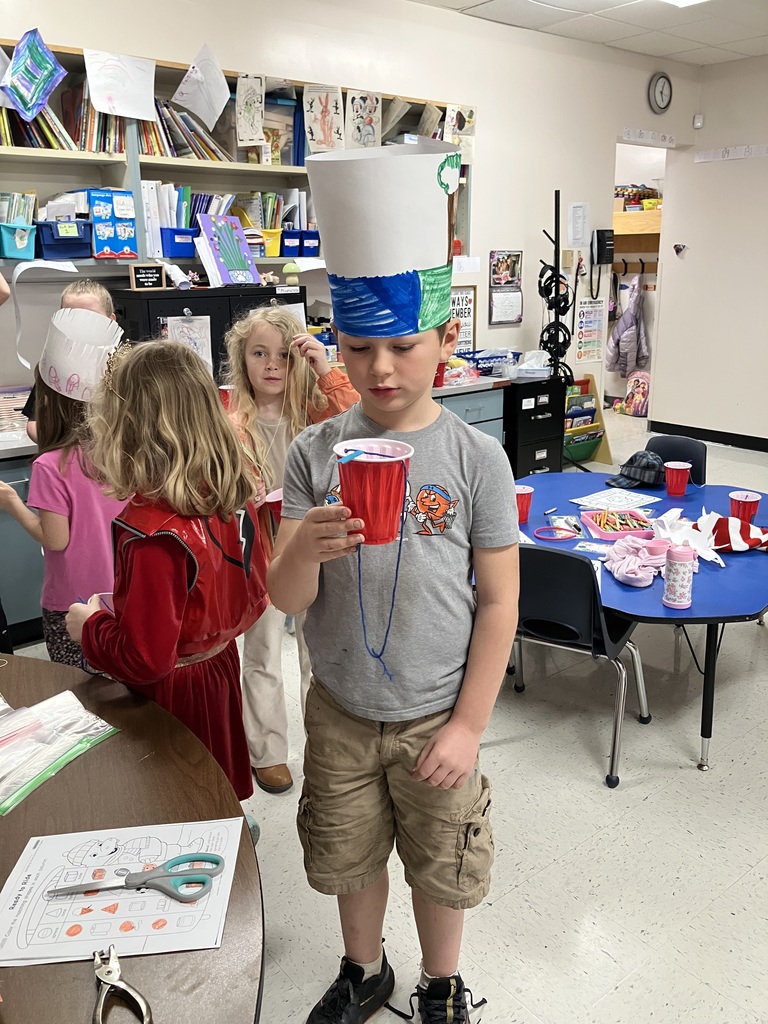  A young boy in a classroom wears a tall, hand-decorated paper crown and holds a red plastic cup with blue yarn threaded through it. Other children and classroom shelves filled with books are visible in the background.  