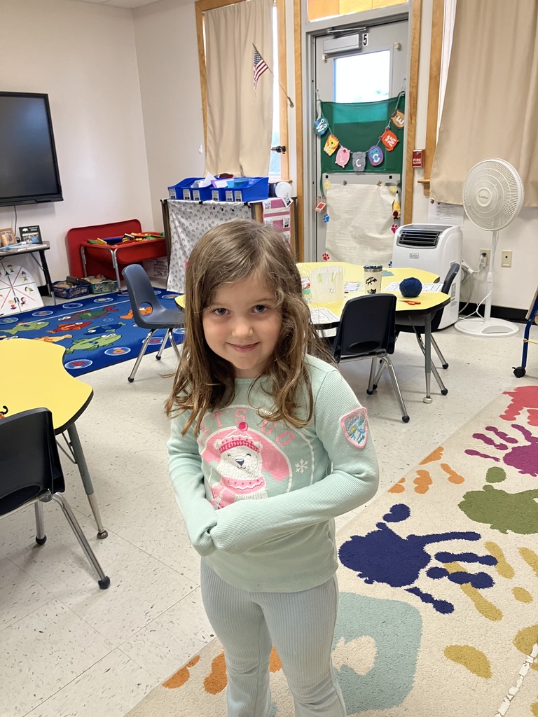  A young girl with wavy brown hair wearing a light green long-sleeved shirt smiles while standing on a colorful rug featuring large handprints. The classroom background includes a yellow table and a door with student decorations.  