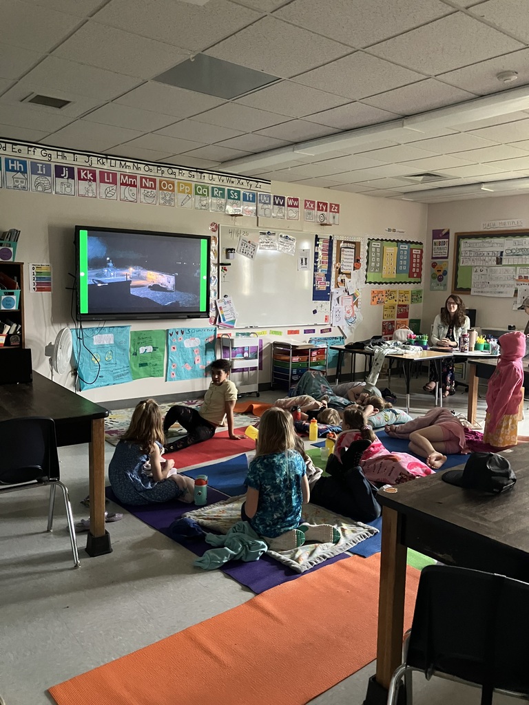: A wide-angle shot of a classroom during a movie screening. Several elementary-aged students are sitting or lying on colorful yoga mats and blankets spread across the floor. A teacher sits at a desk in the background, and a large screen at the front of the room displays a night scene from a film. The room is decorated with educational posters and an alphabet strip. 