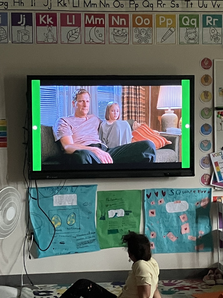  A close-up of a large wall-mounted television in a classroom. The screen shows a scene from a movie featuring an adult man and a young girl sitting together on a couch. Below the TV, colorful student-made posters are taped to the wall, and the top of a student's head is visible in the foreground as they watch. 