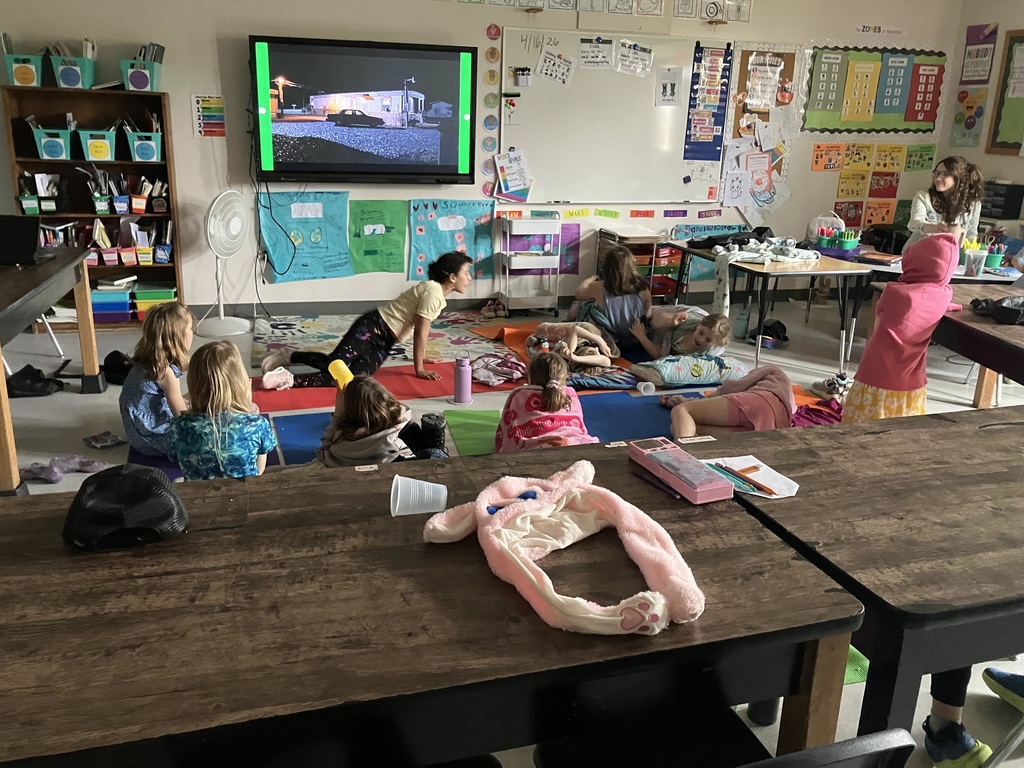  A wide-angle shot of a classroom during a movie screening. Several elementary-aged students are sitting or lying on colorful yoga mats and blankets spread across the floor. A teacher sits at a desk in the background, and a large screen at the front of the room displays a night scene from a film. The room is decorated with educational posters and an alphabet strip.