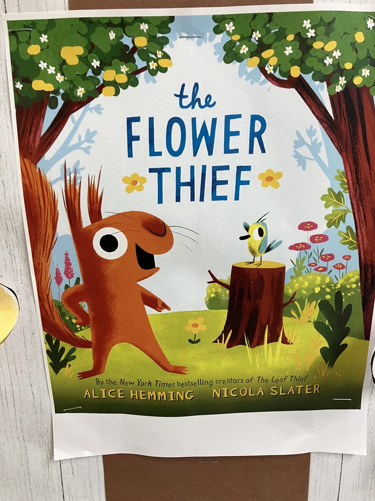  Bulletin Board Display A vibrant, classroom bulletin board themed after the book "The Flower Thief." * Centerpiece: A large, stylized tree made of brown construction paper strips stands in the middle. Hand-shaped cutouts in dark green act as "leaves" on the branches.  Bottom Section: A row of colorful, hand-drawn paper flowers with green stems "grows" along the bottom of the board. The flowers are brightly colored in shades of blue, pink, yellow, and orange.  Decorative Elements: Oversized, decorative paper pencils and bright stars are scattered across the white-plank patterned background.  Book Reference: A printout of the book cover for The Flower Thief by Alice Hemming is pinned directly to the trunk of the paper tree.  Border: The entire board is framed by a colorful border featuring a pattern of vertical, multi-colored pencils.  