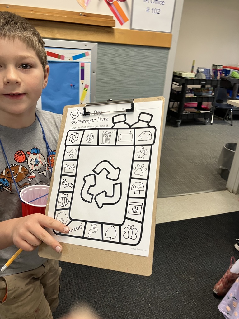 A close-up shot of a young boy holding a clipboard. The clipboard holds a "Earth Day Scavenger Hunt" coloring sheet featuring a large recycling symbol in the center surrounded by icons of nature and sustainable items like flowers, pinecones, and water taps. 