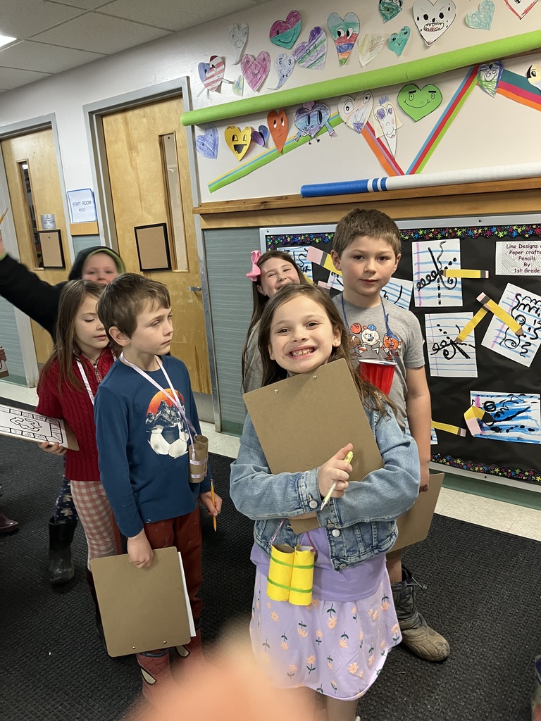  A group of smiling children posing in a school hallway. A girl in a denim jacket and purple dress stands in the center holding a clipboard, with yellow cardboard binoculars hanging from her neck. Behind them is a bulletin board decorated with student artwork and colorful paper hearts.  IMG_3270.jpg: Two children walk toward the camera in a hallway. A girl on the left wears a tall white paper hat and orange jacket, holding a scavenger hunt sheet and green binoculars. A boy behind her peeks through his own cardboard binoculars.
