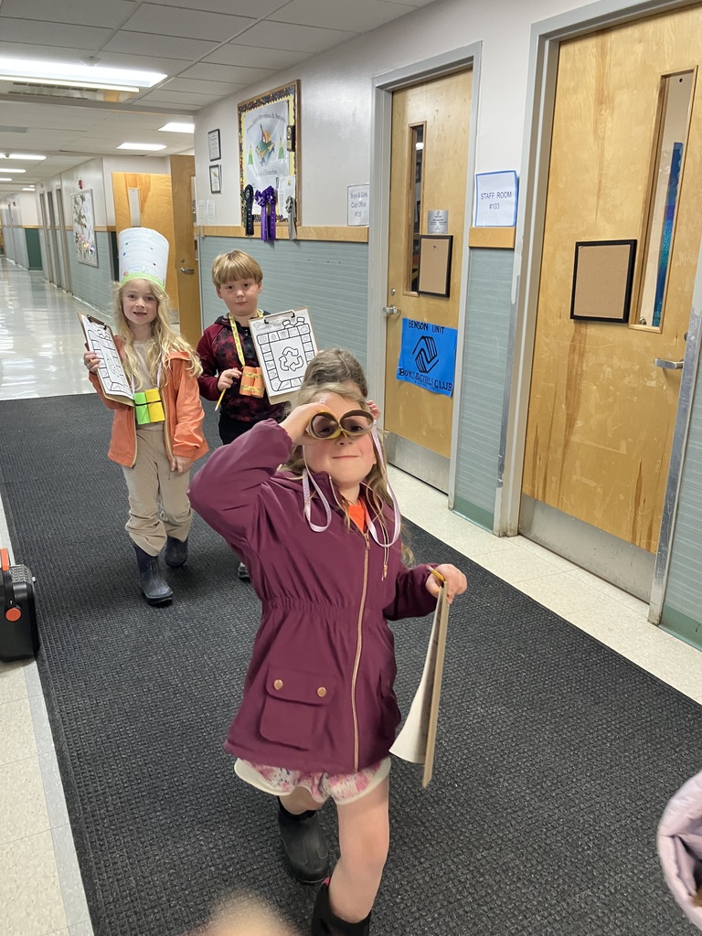  Three children walk down a school hallway. A girl in a purple jacket in the foreground looks through homemade binoculars made from cardboard tubes. Behind her, a girl wears a tall decorated paper hat and a boy holds up his scavenger hunt clipboard. 