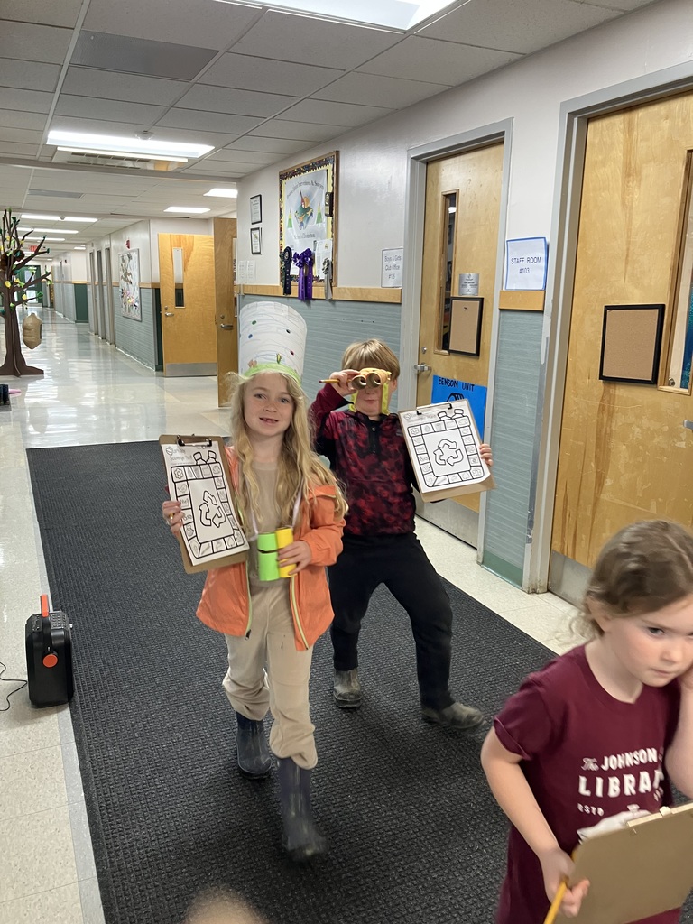  Two children walk toward the camera in a hallway. A girl on the left wears a tall white paper hat and orange jacket, holding a scavenger hunt sheet and green binoculars. A boy behind her peeks through his own cardboard binoculars.