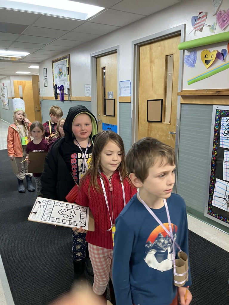 A line of elementary-aged children walking down a school hallway. Several children are holding clipboards with "Earth Day Scavenger Hunt" worksheets. One boy in the foreground wears a blue shirt and has homemade cardboard tube binoculars hanging around his neck.