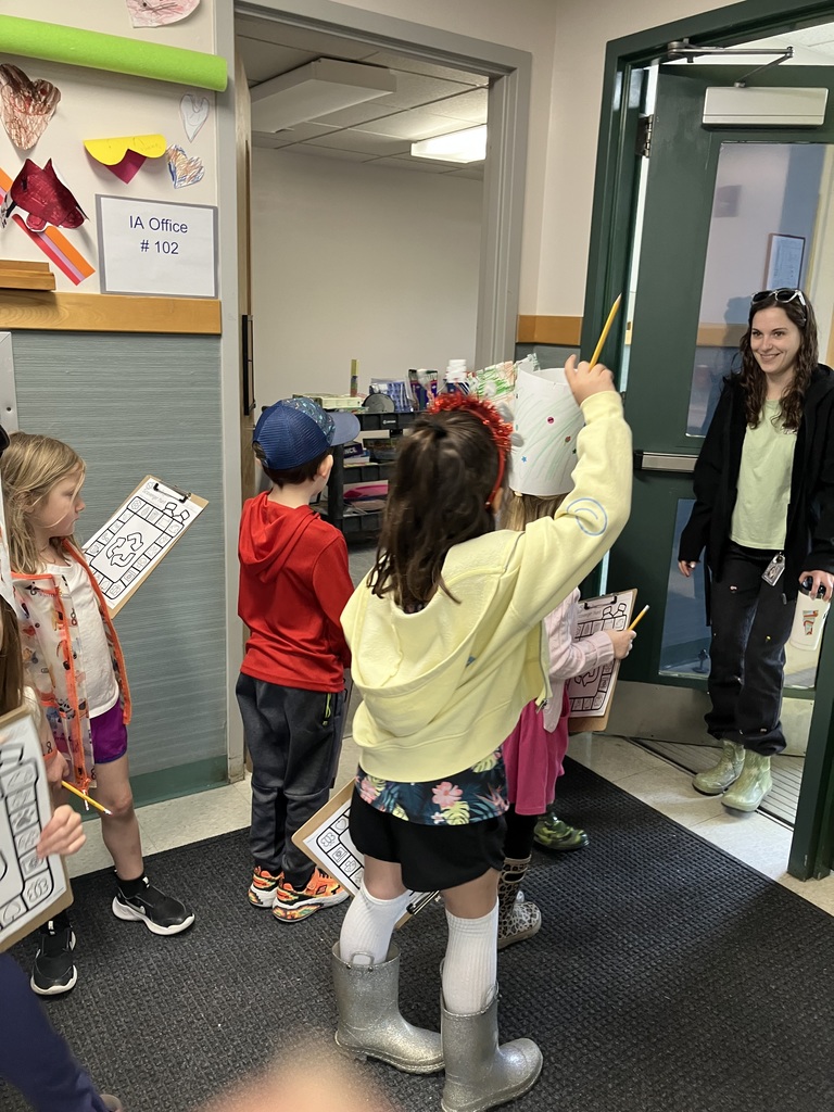  A group of children standing in a hallway near a doorway labeled "IA Office #102." A girl in a yellow hoodie and silver glitter boots holds a pencil up in the air. A female teacher or staff member stands in the doorway smiling at the group.  