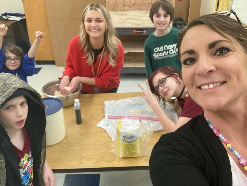 Teacher and several students gather around a table mixing ingredients for a baking activity, smiling and posing for a group selfie in a classroom.