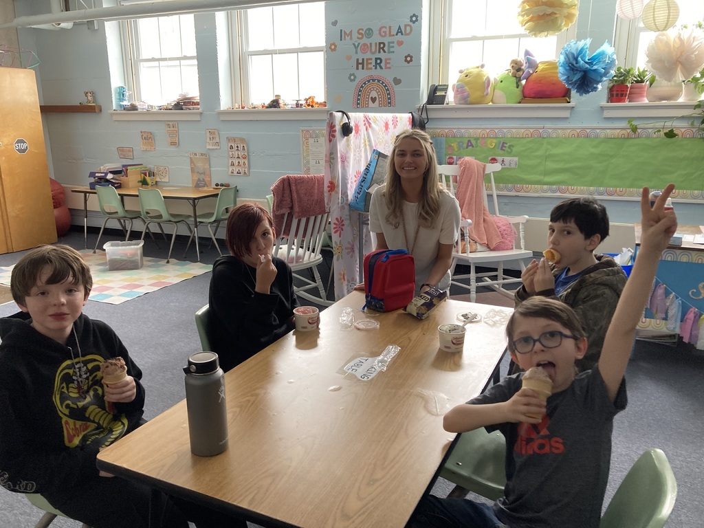 Teacher sits with a group of students around a classroom table as they enjoy ice cream cones and treats during a celebration.