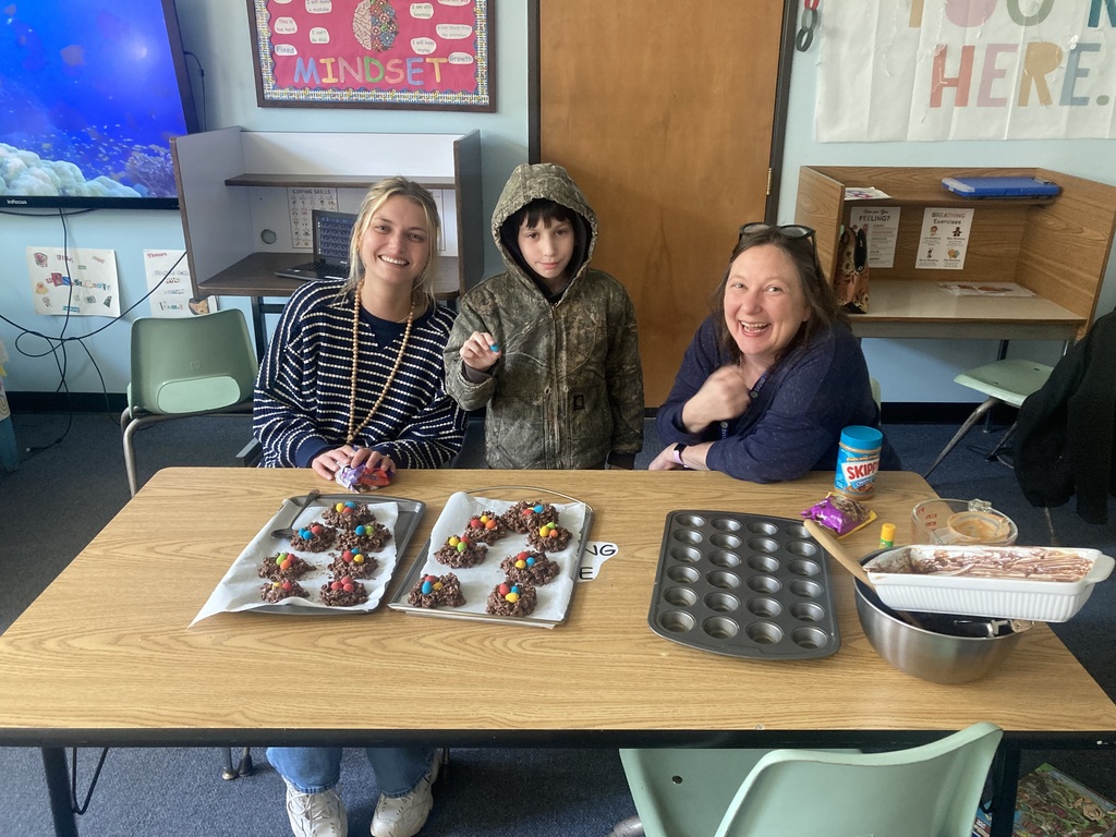 Two staff members and a student smile behind trays of homemade robin’s nest treats topped with candy, with baking supplies on the table in a classroom setting.