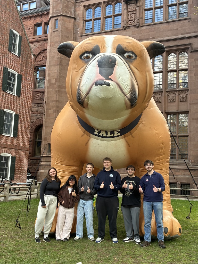 A group of students stand smiling in front of a large inflatable bulldog mascot with a Yale collar, posing outside a historic brick campus building during a college visit.