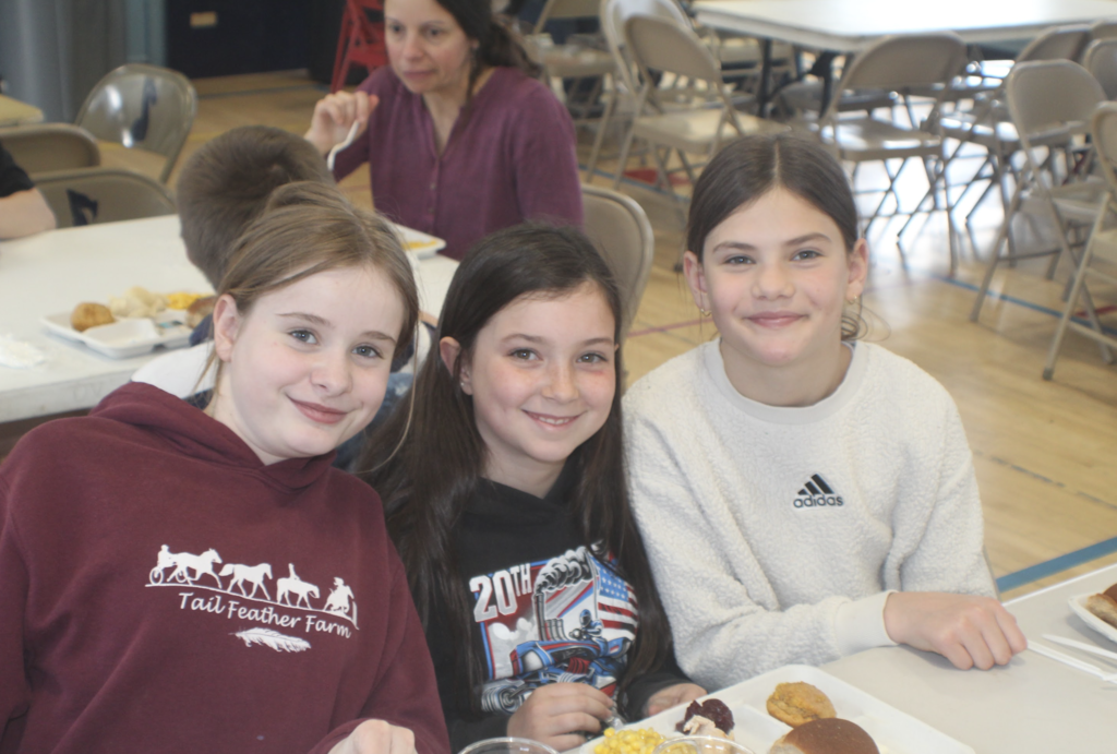 Three girls sit at a cafeteria table with lunch trays, smiling at the camera in a bright, busy dining space.