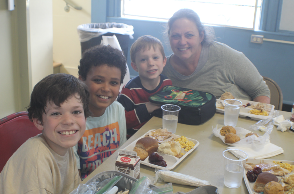 Four children and an adult sit together at a table with lunch trays, smiling at the camera in a crowded cafeteria.