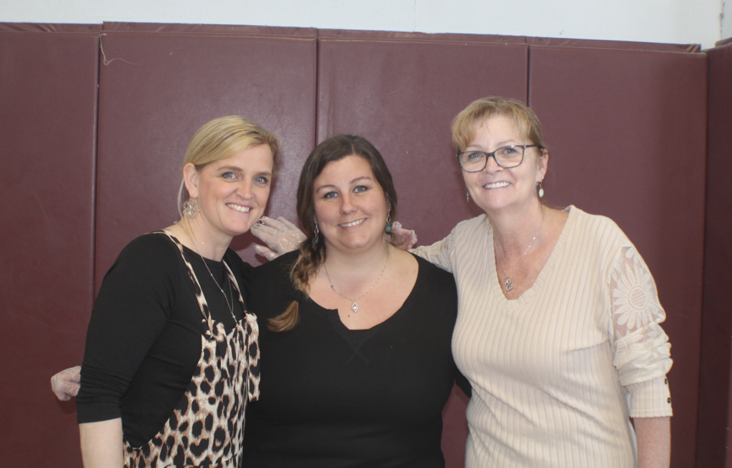 Three adult women stand together smiling in front of a padded gym wall, posing for a group photo.