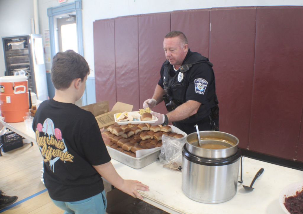 A police officer wearing gloves serves food from a table to a student holding a tray, with rolls and hot food visible.