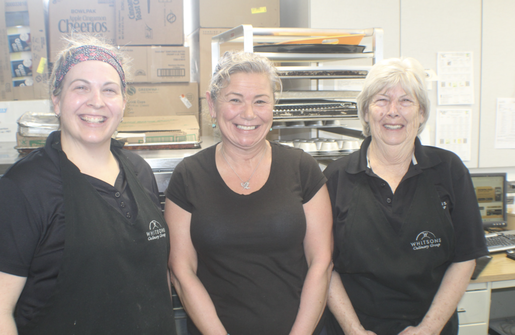 Three cafeteria staff members wearing black shirts and aprons stand together in a kitchen area, smiling at the camera.