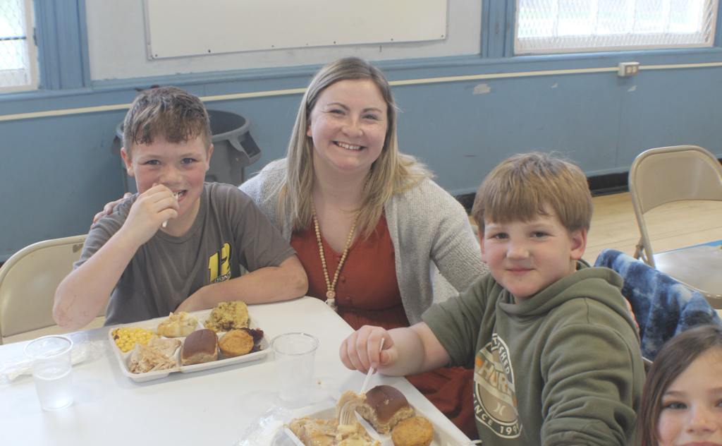 A staff member sits with two boys at a cafeteria table, smiling at the camera while they eat a meal with trays of food.