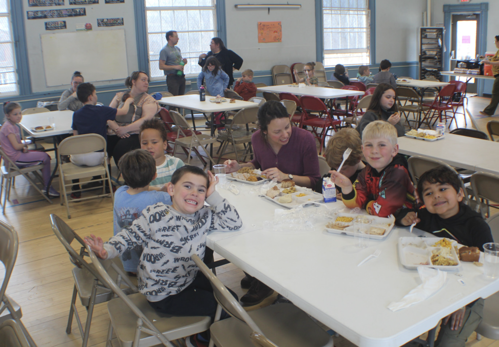 Students and adults sit at long tables in a school cafeteria, eating lunch and talking together. Children smile at the camera while holding trays of food.