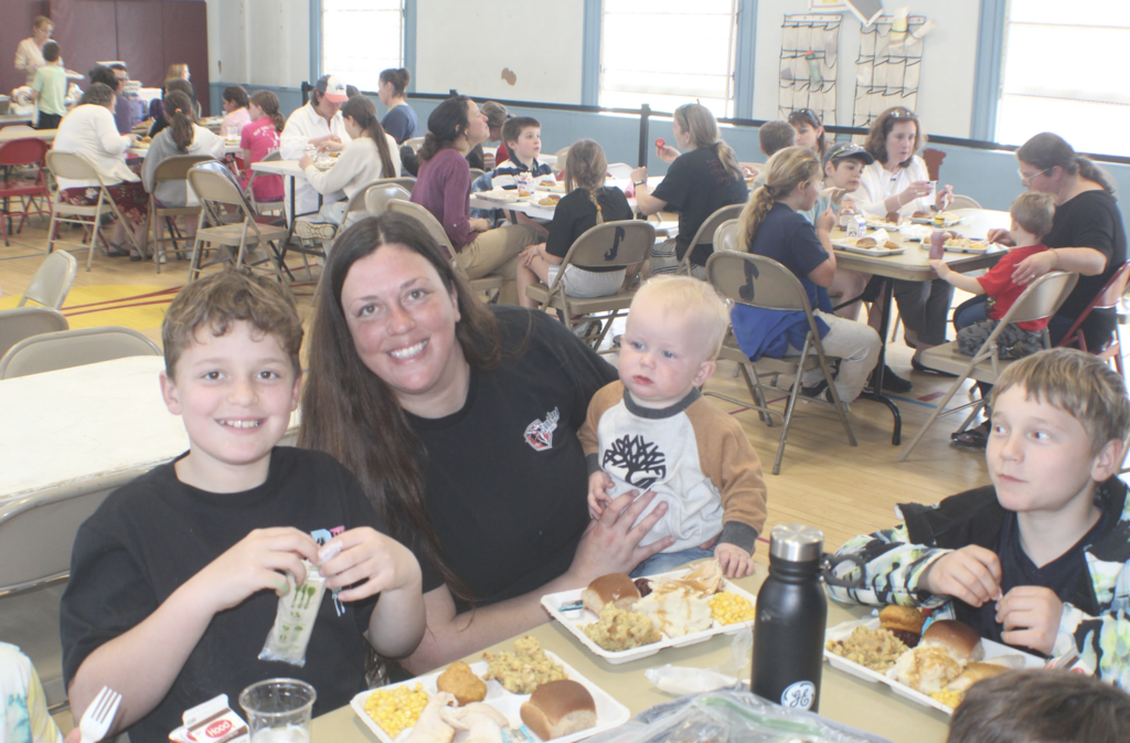 A woman sits with two children and a baby at a cafeteria table, smiling while eating lunch among other families in the background.