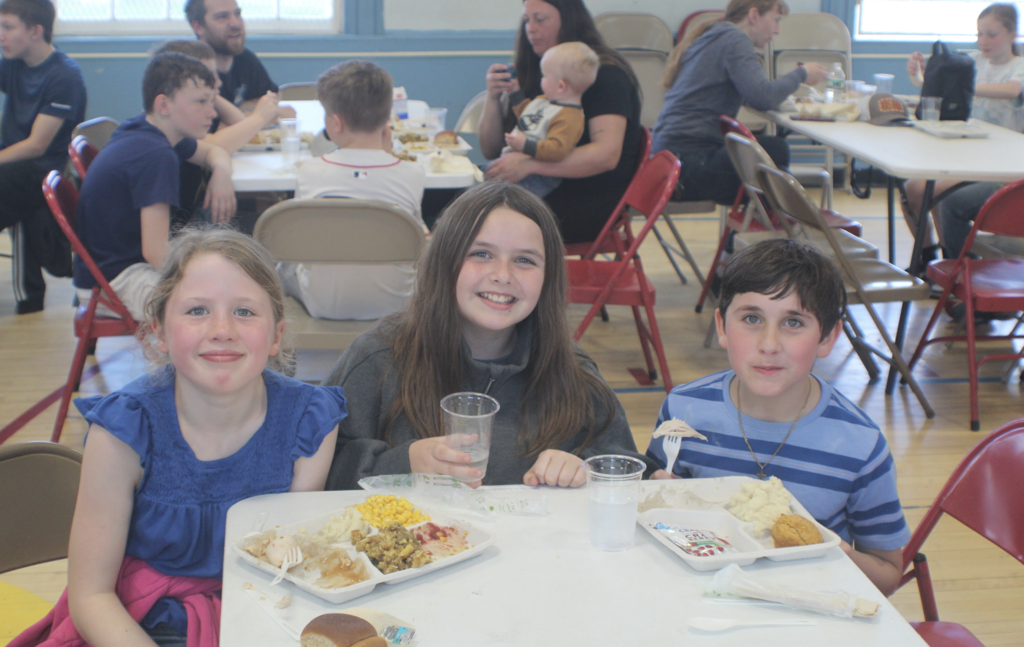Three students sit at a cafeteria table with trays of food, smiling while holding drinks and utensils, with other diners behind them.