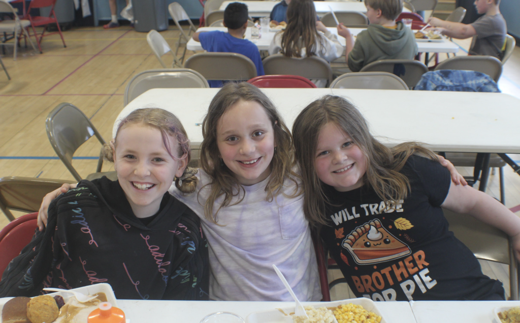 Three elementary-aged girls sit side by side at a cafeteria table, smiling with arms around each other while eating lunch.
