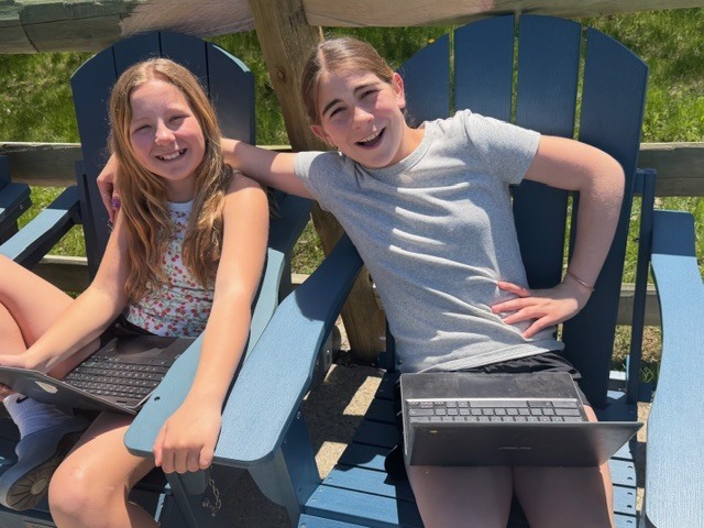 Two students relax in Adirondack chairs outdoors, smiling with laptops on their laps while working together in the sun.