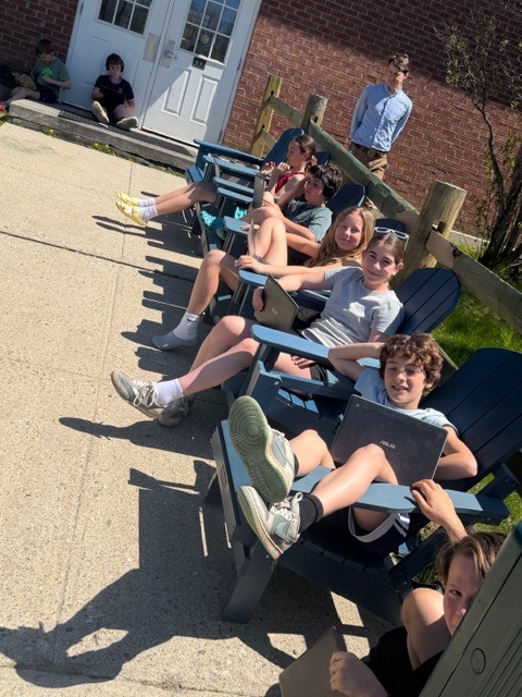 Students sit in outdoor Adirondack chairs along a school building, smiling and using laptops in the sunshine while a teacher stands nearby.