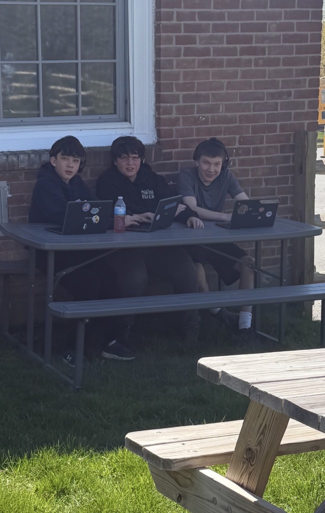 Three students sit at a picnic table outside a brick school building, working on laptops and looking toward the camera.