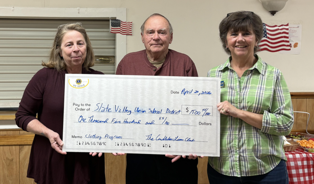 Three adults stand indoors holding a large ceremonial check made out to the Slate Valley Unified School District for $1,500. Two American flags hang on the wall behind them. A table with refreshments is visible to the side.