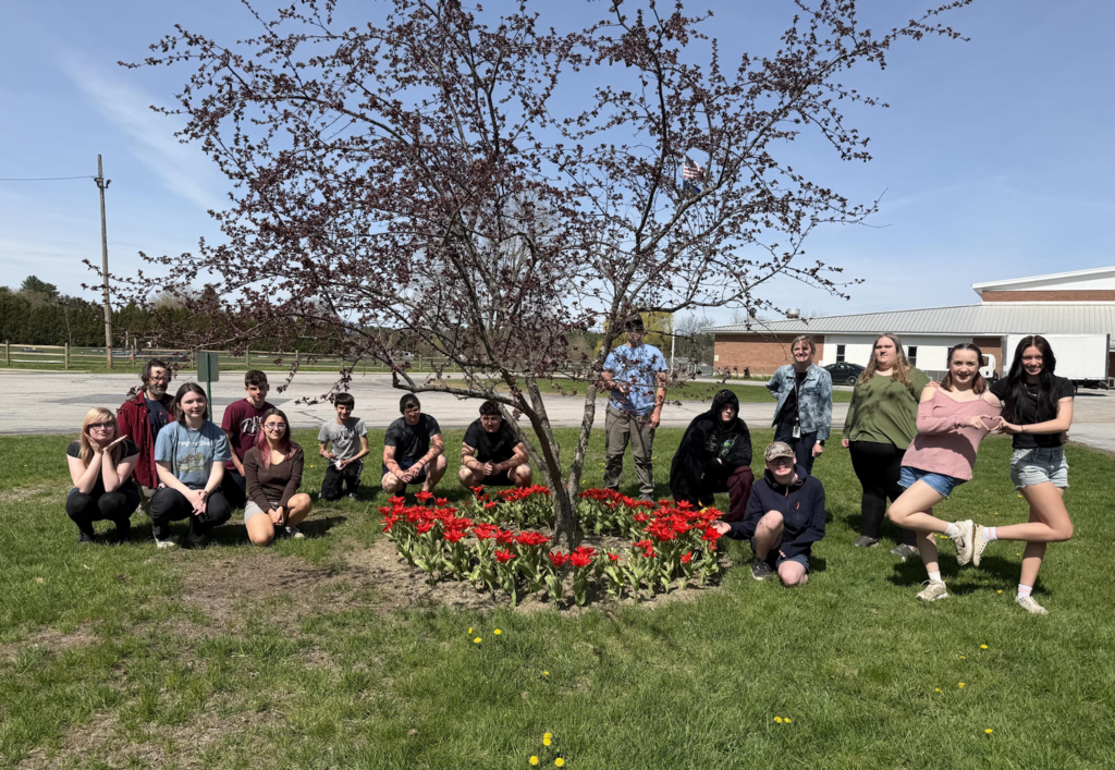 A group of students and staff gather outdoors around a flowering tree and a circular bed of bright red tulips on school grounds, posing together on a sunny day.