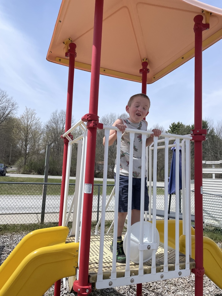 : A young boy in a white dinosaur-patterned tank top and blue cargo shorts stands on a playground platform. He is holding onto white vertical railings with a squinting or strained facial expression. The structure has bright red support poles and a peach-colored canopy. Two yellow slides are visible at the base against a background of trees and a clear blue sky.  