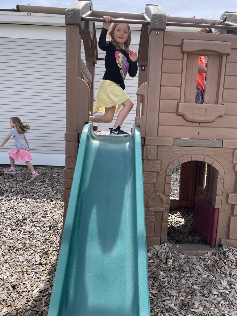  A young girl with long blonde hair, wearing a black long-sleeve shirt with a colorful butterfly graphic and a yellow skirt, prepares to go down a teal-colored slide. She is positioned at the top of a brown, castle-like plastic play structure. In the background, another child in a pink skirt runs across the woodchip-covered ground