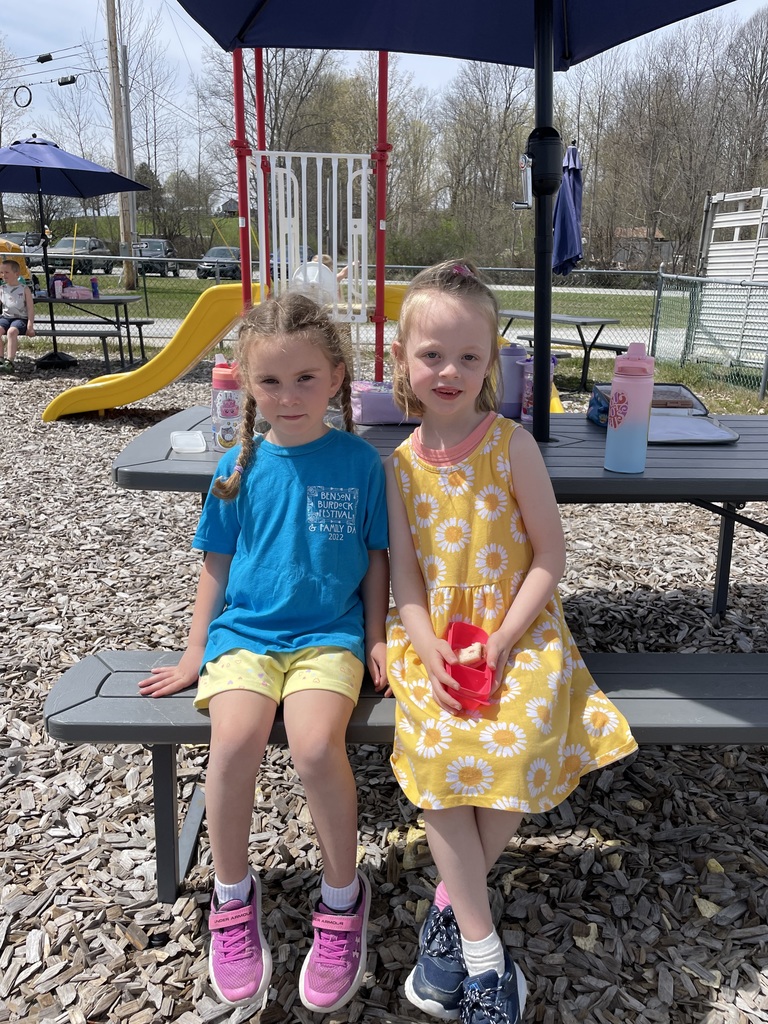  Two young girls sit side-by-side on a gray plastic picnic bench outdoors. The girl on the left wears a blue t-shirt and yellow shorts with her hair in braids; the girl on the right wears a yellow dress with a white daisy pattern and holds a small red snack container. They are sitting under the shade of a blue umbrella on a sunny day, with a playground and woodchips in the
