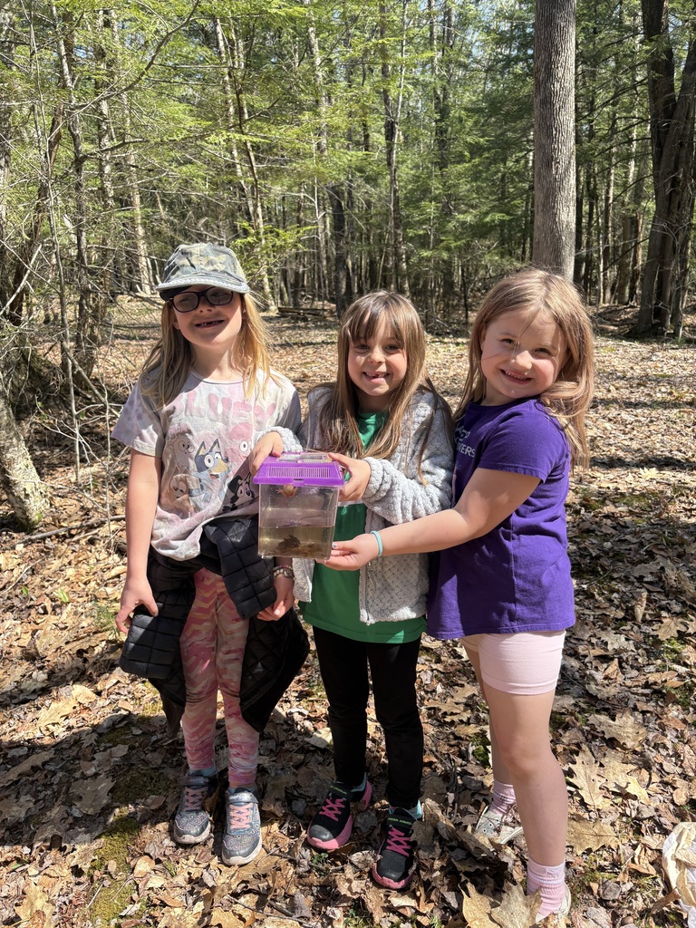 Three young students stand together in a wooded area, smiling and holding a small clear container with a purple lid that contains water and pond creatures they collected during outdoor exploration.