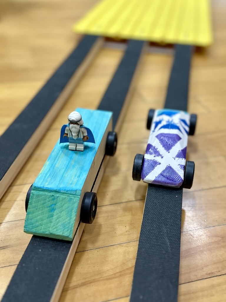Close-up of two pinewood derby cars on a track, one featuring a small figurine and the other painted with a bold purple and white design.