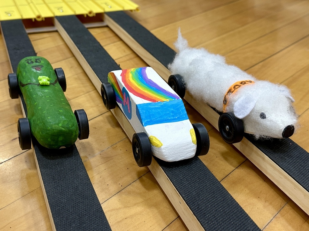 Three pinewood derby cars sit on a wooden race track, including one with a rainbow design and one shaped like a white animal.