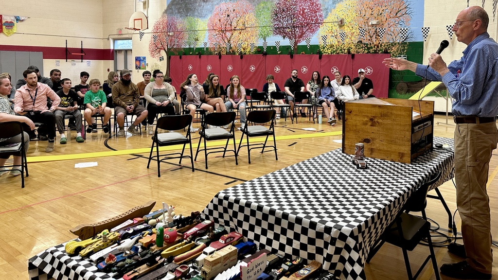 A speaker addresses a seated audience in a school gym during the pinewood derby event, with a table displaying multiple handmade cars.