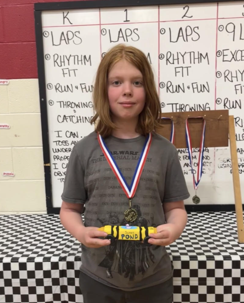 Student with a medal holds a yellow and black pinewood derby car labeled “James Pond,” posing in front of a gym display.