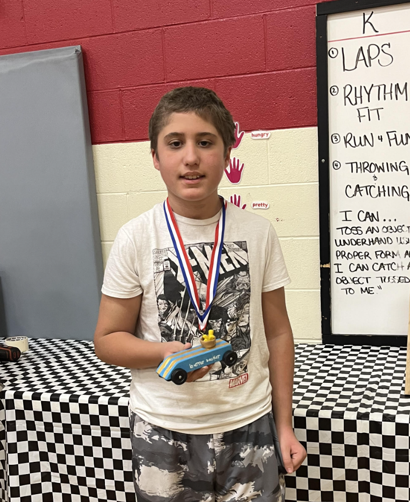 Student wearing a medal holds a blue pinewood derby car while standing in a gymnasium with event signage behind them.