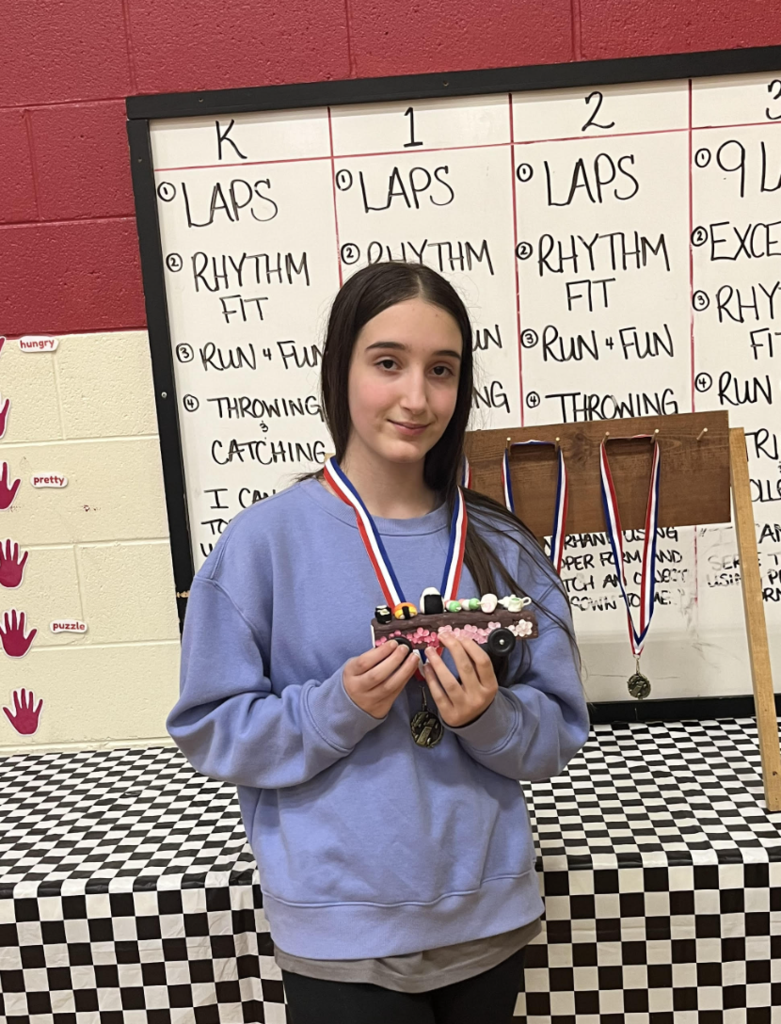 Student wearing a medal holds a decorated pinewood derby car in a school gym, standing in front of a display board and checkered table.