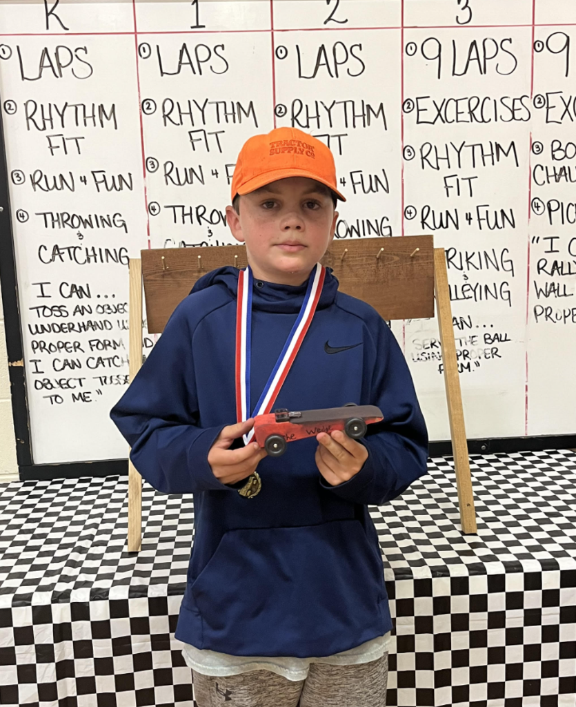 Student in a navy hoodie and orange hat holds a red pinewood derby car labeled “The Wedge,” wearing a medal.