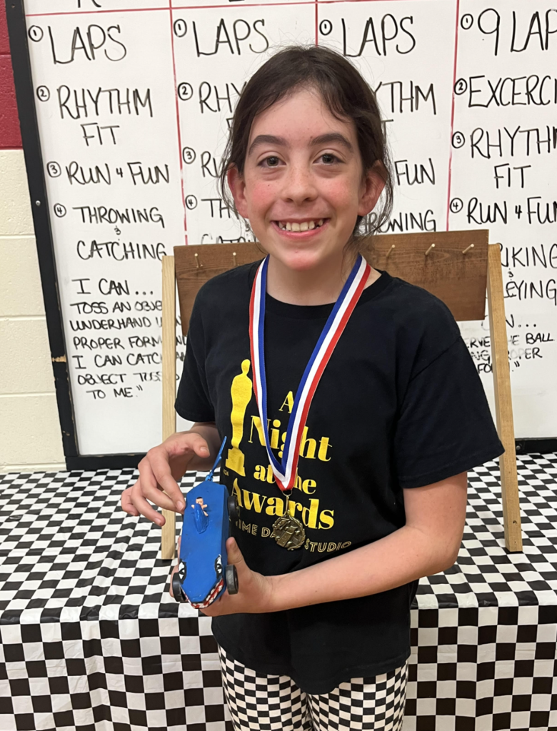 Smiling student wearing a medal holds a blue pinewood derby car, standing in front of a display board in the gym.
