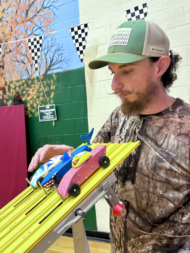 Adult adjusts two colorful pinewood derby cars positioned on a yellow race track, preparing them for a race.