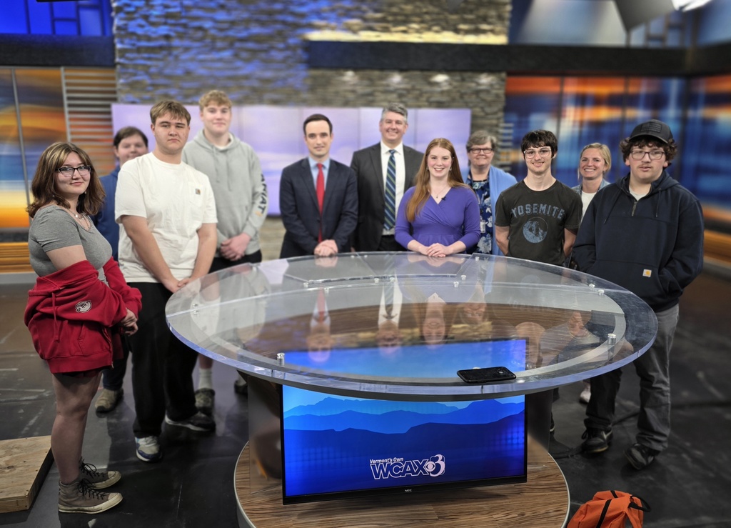 Students and news staff pose together behind a news desk in a television studio with a WCAX Channel 3 display on the front.
