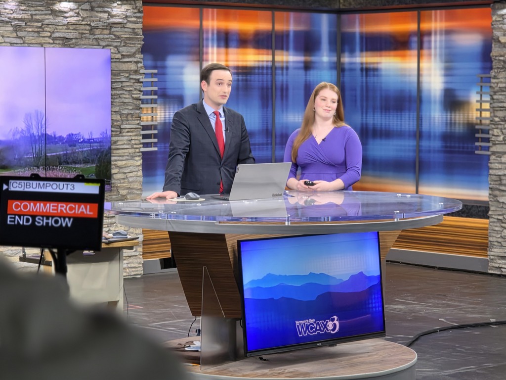 Two news anchors stand behind a desk in a TV studio set, preparing for a broadcast with screens and lighting in the background.