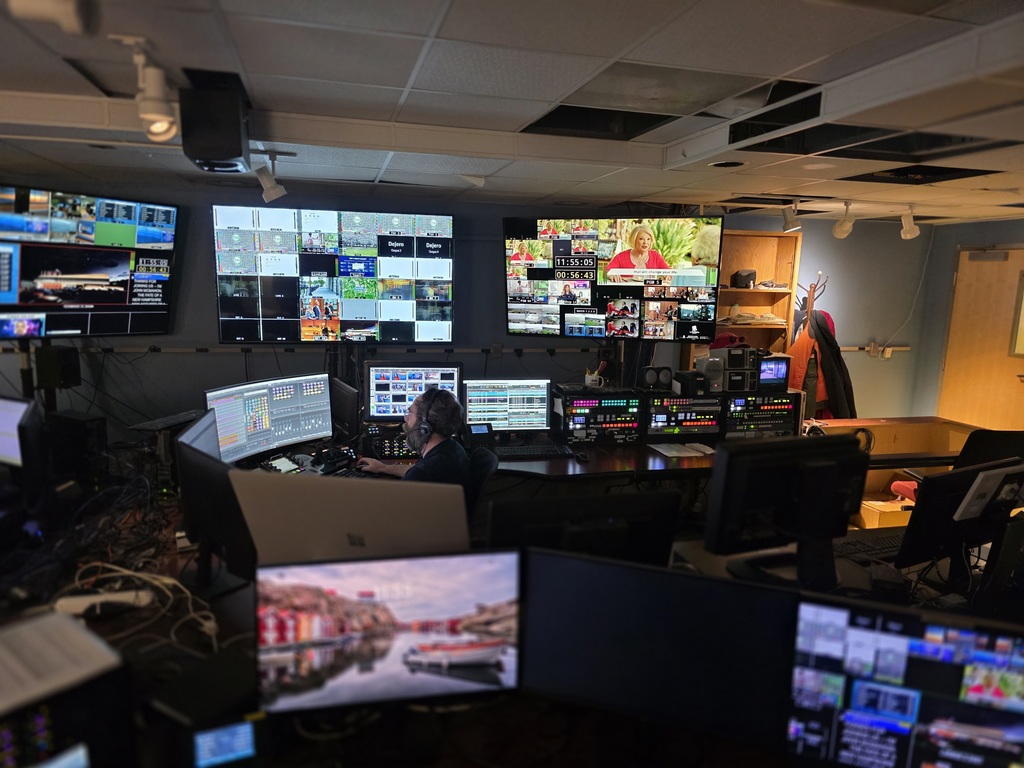 Television control room with a technician wearing a headset operating multiple monitors showing live feeds, graphics, and video controls.