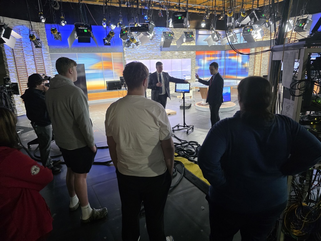 Group of students and adults observe a live TV studio from behind as a presenter gestures near a teleprompter under studio lights and equipment.