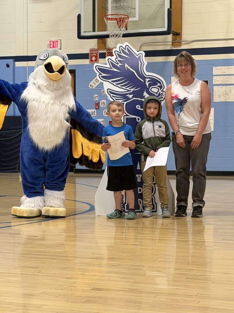 Two young students holding certificates stand in a school gym with an adult and a large bird mascot, posing in front of a basketball hoop and a school eagle display.